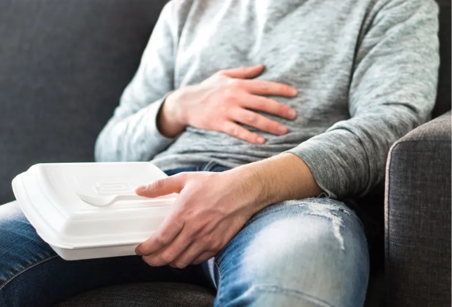 Joven cansado sentado en el sofá, sostiene un táper con una mano y se toca el abdomen con la otra