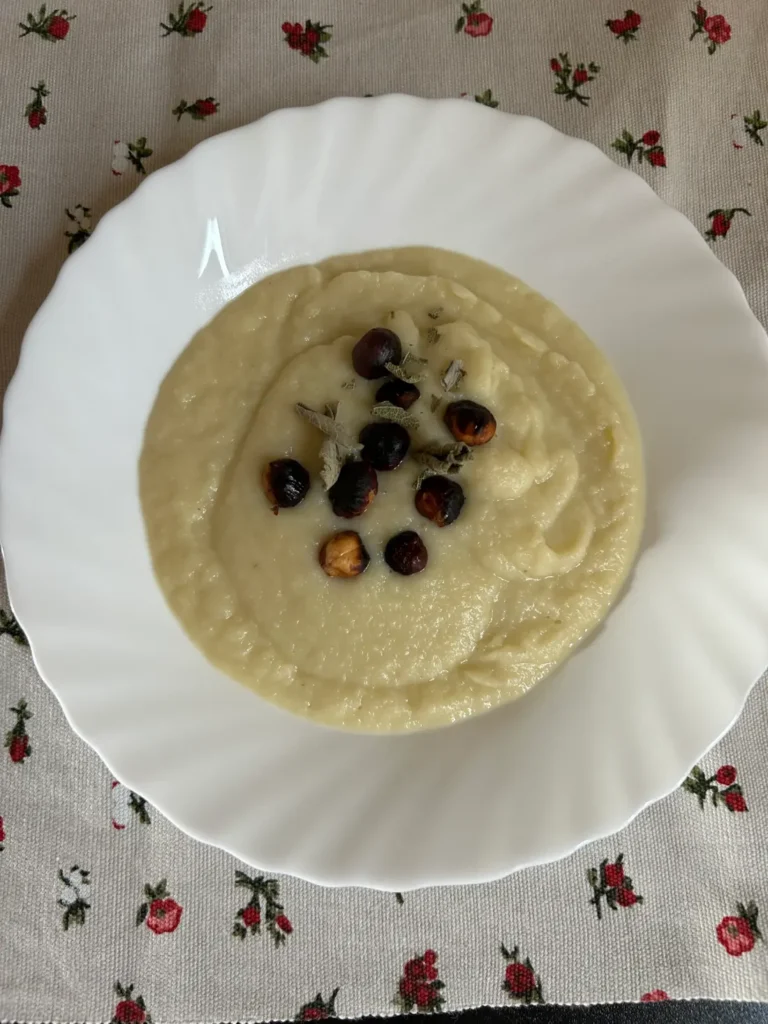 Un plato de sopa crema de color claro, servida en un plato blanco, decorada en el centro con avellanas tostadas y hojas secas de hierbas, sobre un mantel con estampado floral.