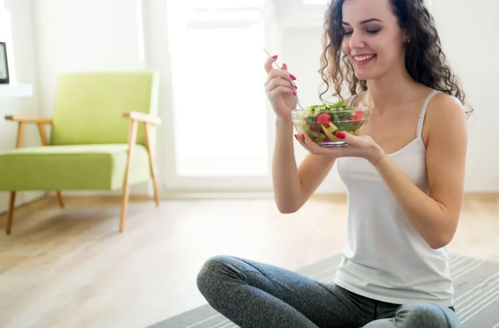 Mujer joven sentada en el suelo sonriendo mientras come una ensalada fresca de un bol, en un ambiente luminoso y relajado.
