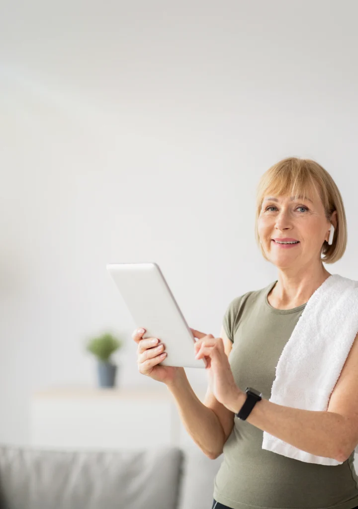 Mujer madura sonriendo mientras sostiene una tablet, con ropa deportiva y una toalla al hombro, en un ambiente luminoso de interior.