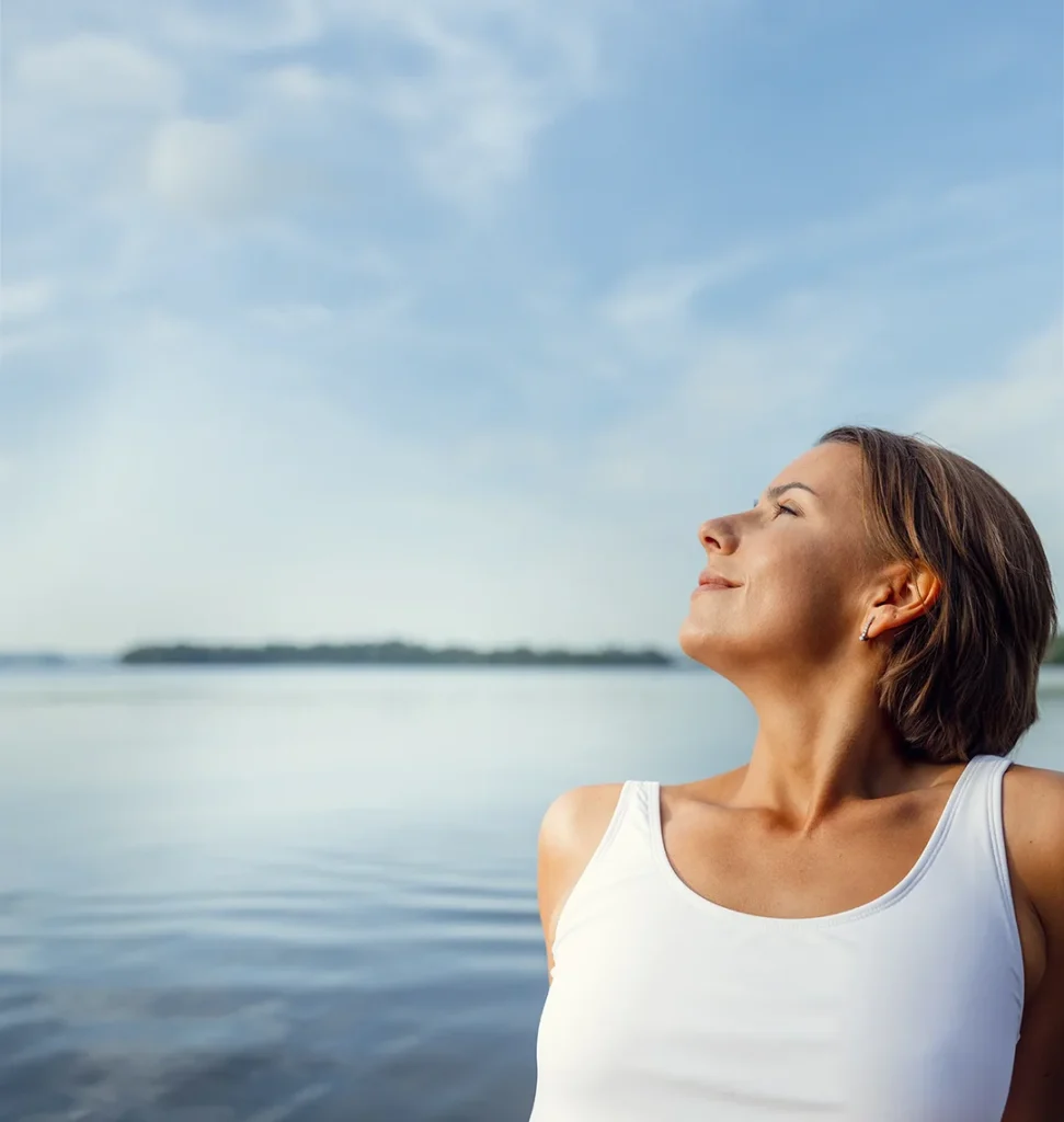 Mujer al aire libre junto al agua, con los ojos cerrados y expresión de bienestar, respirando profundamente en un entorno natural.
