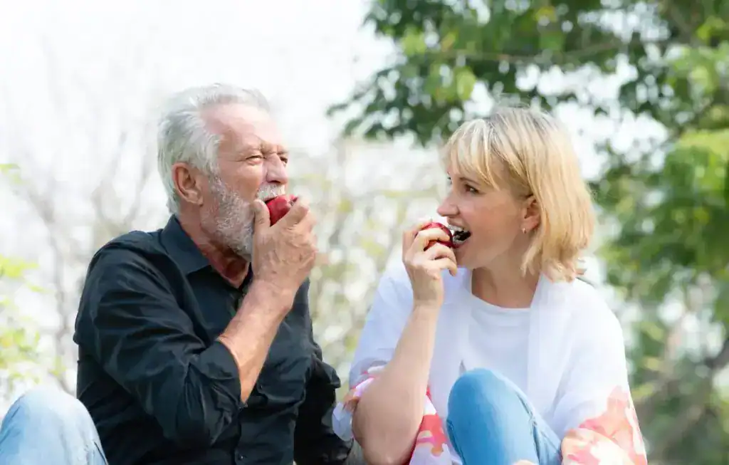Hombre mayor y mujer sonriendo al aire libre mientras comen manzanas rojas, fruta reguladora del tránsito intestinal